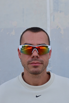 Male model wearing INDY polarized sport sunglasses with a white frame and rainbow mirrored red-orange gradient lenses, standing against a light textured wall.