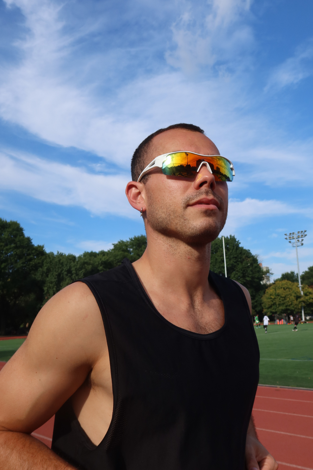 Man wearing reflective Indy Sports sunglasses on a sports field with trees and sky in the background